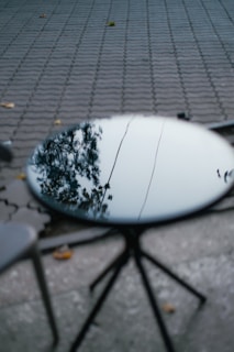 A round table with a reflective surface shows the reflection of nearby trees and a clear sky. The table is positioned on a paved area with a pattern of interlocking bricks. A few scattered leaves are visible around the table, suggesting an outdoor setting.