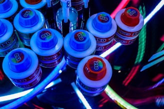 Several small, round containers with colorful screw-on lids labeled with numbers like 150, 250, and 500 are arranged in a cluster. The scene is lit by vibrant, multicolored LED lights, creating a visually dynamic and somewhat futuristic atmosphere. Some tools with white handles are also partially visible among the containers.
