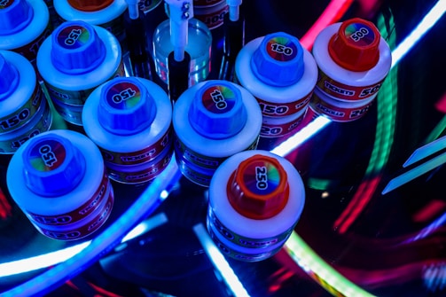 Several small, round containers with colorful screw-on lids labeled with numbers like 150, 250, and 500 are arranged in a cluster. The scene is lit by vibrant, multicolored LED lights, creating a visually dynamic and somewhat futuristic atmosphere. Some tools with white handles are also partially visible among the containers.