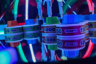 Stacks of tickets are arranged on a reflective surface, featuring various numerical values. Vibrant neon lights in red, blue, and green create a lively, arcade-like atmosphere, casting colorful reflections.