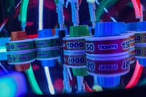 Stacks of tickets are arranged on a reflective surface, featuring various numerical values. Vibrant neon lights in red, blue, and green create a lively, arcade-like atmosphere, casting colorful reflections.