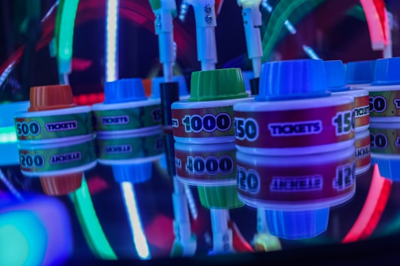 Stacks of tickets are arranged on a reflective surface, featuring various numerical values. Vibrant neon lights in red, blue, and green create a lively, arcade-like atmosphere, casting colorful reflections.