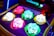 A colorful arcade game machine featuring illuminated clown heads with different expressions. The clowns are embedded in a whack-a-mole style setup, with a mallet to the side suggesting gameplay. The heads are brightly lit in various colors including pink, green, and blue, with exaggerated facial features.