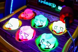 A colorful arcade game machine featuring illuminated clown heads with different expressions. The clowns are embedded in a whack-a-mole style setup, with a mallet to the side suggesting gameplay. The heads are brightly lit in various colors including pink, green, and blue, with exaggerated facial features.