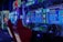 A vibrant arcade environment with several illuminated gaming machines. The image features arcade racing games with visible steering wheels, colorful control panels, and screens displaying game instructions. Bright LED lights in various colors illuminate the machines and surrounding area.