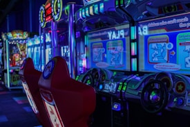 A vibrant arcade environment with several illuminated gaming machines. The image features arcade racing games with visible steering wheels, colorful control panels, and screens displaying game instructions. Bright LED lights in various colors illuminate the machines and surrounding area.