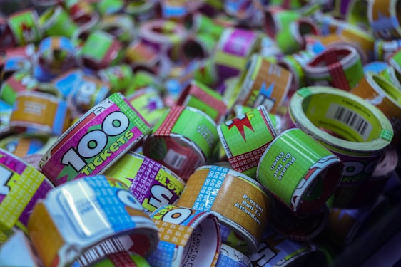 A collection of colorful arcade tickets, predominantly green, pink, and blue, rolled up and stacked together. The tickets display various designs and text, including one that prominently shows '100 tickets' in bold green numbers on a pink background.
