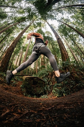 A person wearing athletic attire is captured mid-stride while running through a lush forest with towering trees. The perspective is from below, emphasizing movement and the height of the trees.