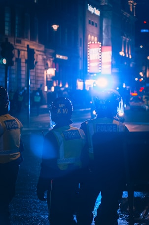 Police officers wearing reflective vests stand in a dimly lit urban setting at night. The scene is illuminated by bright blue lights, creating a moody atmosphere. Buildings in the background have glowing signs and a crowd can be seen in the distance.