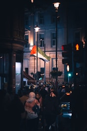 A crowd of people is gathered in a city street at night, near a traffic light and several street lamps. A large flag with red, white, and green colors is prominently displayed. The background includes a tall building with several windows lit up.