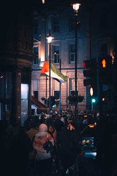 A crowd of people is gathered in a city street at night, near a traffic light and several street lamps. A large flag with red, white, and green colors is prominently displayed. The background includes a tall building with several windows lit up.