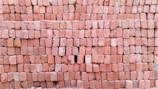A large stack of reddish-brown bricks neatly arranged in a rectangular pattern. The surface of the bricks is textured, and some bricks have slight variations in color. There is a small gap in the arrangement where a few bricks are missing.