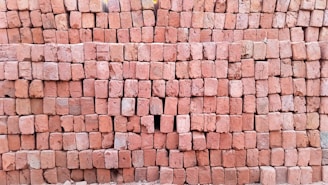 A large stack of reddish-brown bricks neatly arranged in a rectangular pattern. The surface of the bricks is textured, and some bricks have slight variations in color. There is a small gap in the arrangement where a few bricks are missing.
