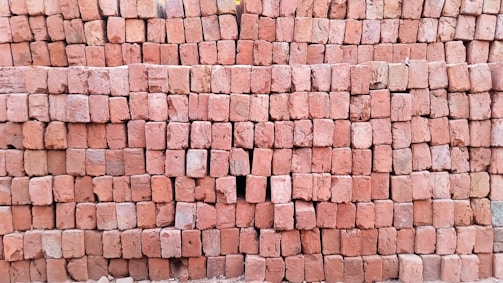 A large stack of reddish-brown bricks neatly arranged in a rectangular pattern. The surface of the bricks is textured, and some bricks have slight variations in color. There is a small gap in the arrangement where a few bricks are missing.