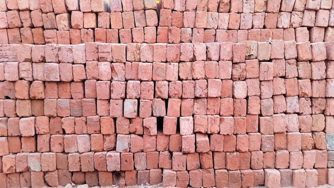A large stack of reddish-brown bricks neatly arranged in a rectangular pattern. The surface of the bricks is textured, and some bricks have slight variations in color. There is a small gap in the arrangement where a few bricks are missing.
