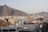 An urban landscape with densely packed buildings sits against a backdrop of brown, rocky mountains. The architecture is a mix of modern and traditional styles, with a large number of boxy, flat-roofed structures. A few taller buildings rise above the others, and there's a visible presence of satellite dishes and water tanks on the rooftops. A van and some debris are seen on the foreground roof, with a clear view of the surrounding cityscape.