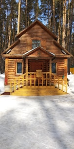 A wooden log cabin with intricate carvings stands in a snowy forest. The building features decorative elements around the door and windows, with a wooden ramp leading to the entrance. Tall pine trees surround the structure, creating a serene and rustic atmosphere.