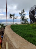 A lush green lawn with a curved stone border overlooks a scenic view. In the background, two small trees stand against a cloudy sky. To the right, a modern, spiral staircase with a dark metal railing ascends, adding an architectural element.