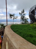 A lush green lawn with a curved stone border overlooks a scenic view. In the background, two small trees stand against a cloudy sky. To the right, a modern, spiral staircase with a dark metal railing ascends, adding an architectural element.