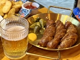 A glass of beer with intricate patterns on the glass is placed beside a serving of sliced sausages arranged on a brass pan. Accompaniments include small pickles and a dollop of mustard. In the background, a colorful dish with potato wedges and a small portion of ketchup can be seen.