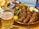 A glass of beer with intricate patterns on the glass is placed beside a serving of sliced sausages arranged on a brass pan. Accompaniments include small pickles and a dollop of mustard. In the background, a colorful dish with potato wedges and a small portion of ketchup can be seen.