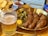 A glass of beer with intricate patterns on the glass is placed beside a serving of sliced sausages arranged on a brass pan. Accompaniments include small pickles and a dollop of mustard. In the background, a colorful dish with potato wedges and a small portion of ketchup can be seen.