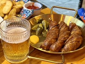 A glass of beer with intricate patterns on the glass is placed beside a serving of sliced sausages arranged on a brass pan. Accompaniments include small pickles and a dollop of mustard. In the background, a colorful dish with potato wedges and a small portion of ketchup can be seen.