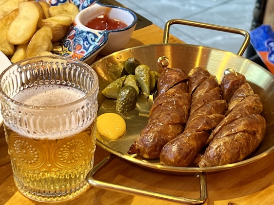 A glass of beer with intricate patterns on the glass is placed beside a serving of sliced sausages arranged on a brass pan. Accompaniments include small pickles and a dollop of mustard. In the background, a colorful dish with potato wedges and a small portion of ketchup can be seen.