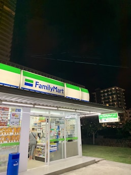 A FamilyMart convenience store is brightly lit at night, with a person standing at the counter inside. The store has glass doors displaying various advertisements and product posters. The store signage is illuminated with green and blue colors. In the background, there are tall, dimly-lit buildings and a dark sky.