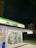 A FamilyMart convenience store is brightly lit at night, with a person standing at the counter inside. The store has glass doors displaying various advertisements and product posters. The store signage is illuminated with green and blue colors. In the background, there are tall, dimly-lit buildings and a dark sky.
