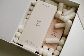 Two rectangular boxes labeled 'SALT & STONE' are nestled inside a white cardboard box filled with white packing peanuts. One box is larger and beige, while the other is slightly smaller and pink. The larger box is labeled as 'Bergamot & Hinoki Body Wash' and the smaller one as 'Natural Deodorant Gel'.
