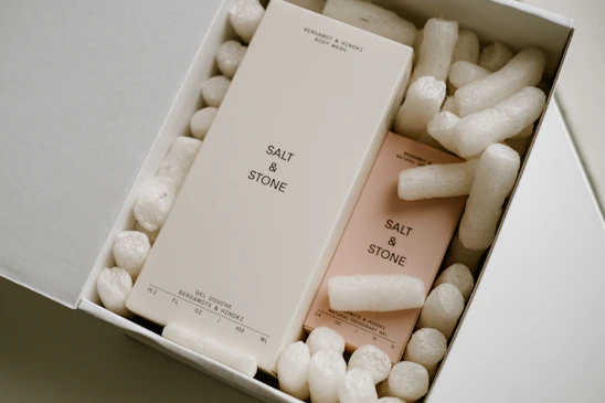 Two rectangular boxes labeled 'SALT & STONE' are nestled inside a white cardboard box filled with white packing peanuts. One box is larger and beige, while the other is slightly smaller and pink. The larger box is labeled as 'Bergamot & Hinoki Body Wash' and the smaller one as 'Natural Deodorant Gel'.
