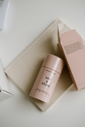A cylindrical container of natural deodorant gel with the brand name 'Salt & Stone' lies on a beige surface next to its matching open box. The container has a minimalist design in a light pink shade with black text.
