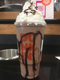 A large iced beverage topped with whipped cream and chocolate drizzle, served in a clear plastic cup with a lid. The cup features the logo of a popular coffee and donut chain in orange and pink lettering. In the background, there is indoor signage, including a QR code for a mobile app.