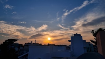 The image captures a vibrant sunset over an urban skyline, with an array of clouds in the sky reflecting shades of orange and gold. Silhouettes of buildings and structures are seen in the foreground against the colorful backdrop of the setting sun.