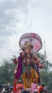 A large statue of a deity, adorned with ornate decorations such as garlands, colorful drapery, and a parasol overhead. The statue is depicted holding a bow and arrow, with a serene expression on its face. Several people dressed in festive attire stand in front, possibly participating in a procession. Trees and a cloudy sky form the background.