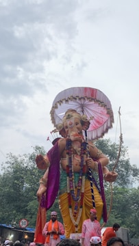 A large statue of a deity, adorned with ornate decorations such as garlands, colorful drapery, and a parasol overhead. The statue is depicted holding a bow and arrow, with a serene expression on its face. Several people dressed in festive attire stand in front, possibly participating in a procession. Trees and a cloudy sky form the background.