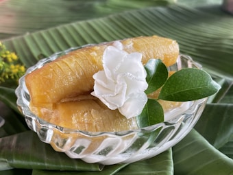 A glass dish contains caramelized bananas topped with a decorative white flower and green leaves, set on a banana leaf backdrop.