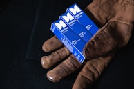 A pair of brown leather gloves holds three blue boxes labeled 'Elemem, Oils for Men'. Each box has different labels: Sleep, Refuel, and Stress. The background is a dark fabric, providing contrast to the objects.