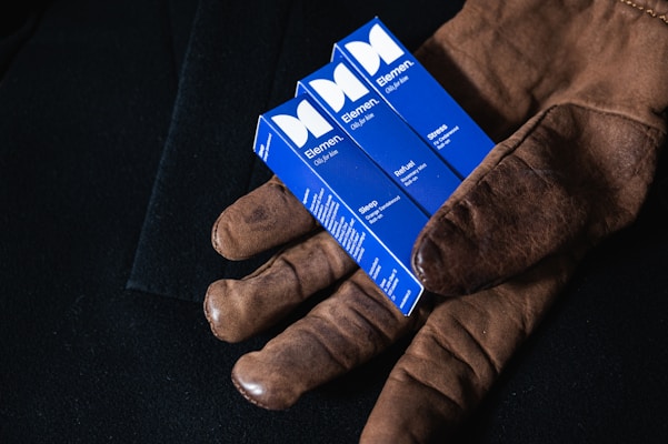A pair of brown leather gloves holds three blue boxes labeled 'Elemem, Oils for Men'. Each box has different labels: Sleep, Refuel, and Stress. The background is a dark fabric, providing contrast to the objects.
