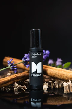 A black roll-on bottle labeled 'Funky Feet' and 'Tea Tree Roll-on' with the brand name 'Elemen'. It is surrounded by cinnamon sticks and purple flowers against a dark background.