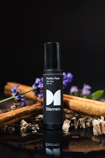 A black roll-on bottle labeled 'Funky Feet' and 'Tea Tree Roll-on' with the brand name 'Elemen'. It is surrounded by cinnamon sticks and purple flowers against a dark background.