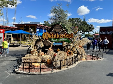 A festive outdoor setting featuring an Oktoberfest food festival sign adorned with various fall-themed decorations, such as hay bales, pumpkins, and corn stalks. Several people are walking around the area, and colorful buildings and structures, including a dining area with blue umbrellas and picnic tables, are visible in the background. The sky is clear with a few scattered clouds, indicating a sunny day.