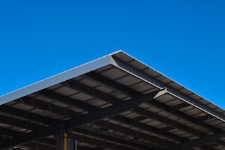 A large, angular metal roof structure set against a clear, vibrant blue sky. The roof features a grid-like pattern of beams and panels.