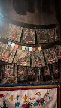 A series of traditional thangka paintings are hung on a dark, worn wall. Each artwork displays intricate religious or spiritual figures with a predominant use of earth tones. Below them are colorful prayer flags featuring multiple hues such as blue, red, yellow, and green. The lower part of the image shows a richly adorned textile featuring vibrant patterns, symbols, and more depictions of spiritual iconography.