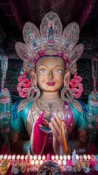 A large, intricately crafted statue of a deity adorned with a detailed and colorful headdress. The statue features rich garments, elaborate jewelry, and vibrant colors, conveying a sense of divinity and reverence. In front, small offerings and a row of lit candles enhance the spiritual ambiance.