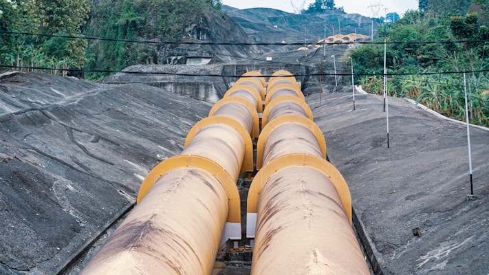 Large yellow pipes are positioned in a parallel pattern across a hilly landscape with vegetation on either side. The pipes stretch into the distance, traversing a series of small hills. Power lines are visible above, and the area is surrounded by greenery, suggesting a rural or industrial setting.
