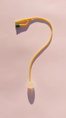 A yellow rubber tubing, resembling a question mark shape, is set against a soft pink background. The top end of the tubing features a green wrapping or band, and there is a white circular object at the lower part.