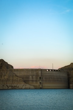 An aerial view of a large dam holding back a vast reservoir under a clear sky.
