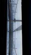 A view of a metal gratin with a person walking on it, seen from below. The structure consists of a grid of metal bars, allowing light to pass through and casting shadows onto the surface below. The scene has a geometric, industrial feel.